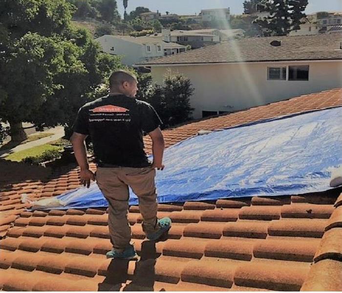 Technician on top of a roof, with the blue tarp covering the spanish tile. Overlooking the neighborhood.